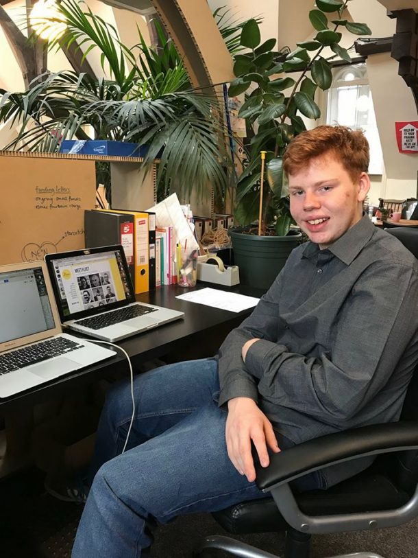 照片中，安德鲁坐在办公椅上，面前是两台笔记本电脑，周围是室内植物。