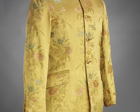 尼赫鲁式夹克，特点是立领和长线条剪裁。这件夹克是由合成织锦织物制成的，有黄色的地面和蓝色、粉色和绿色的花卉图案。上衣从颈部到腰部有四个自盖扣。